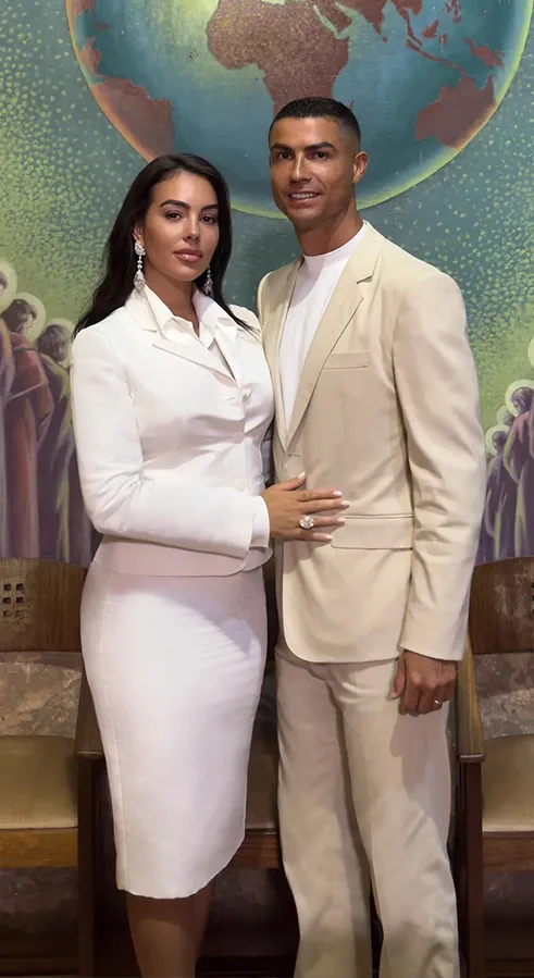 Cristiano Ronaldo and Georgina Rodríguez posing together in elegant white outfits in front of a globe-themed mural.
